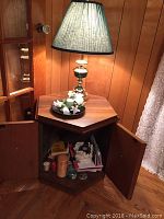 Six-sided side table with wood finish, closed doors open to reveal storage, lamp on top illuminated.