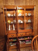 Photo showing inside a china cabinet with open glass doors. Displayed are several sets of tea cups and saucers in floral and solid yellow patterns, glassware, and two white ceramic figurines on middle shelves. Below, three small drawers and open lower shelves hold greeting cards and paper items.