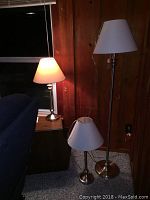 Photo showing all three lamps together: one floor lamp behind two table lamps with lit table lamp illuminating wooden wall.