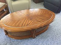 Full view of the wooden oval coffee table with patterned top and cane shelf base on carpeted floor