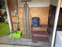 Full view of ironing board, electric iron on wooden shelf, spin mop bucket and floral trash can on metal shelf in a basement area