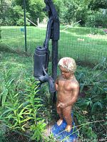 Full view of the cement boy statue standing beside the tall black water pump outdoors in garden setting