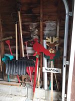 Wooden-handled snow shovels, red broom, and a wooden holiday-themed decorative welcome sign with reindeer and cardinal in a garage corner with wooden walls.