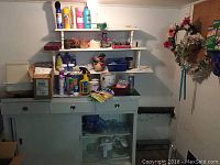 Wide view of cabinet and shelves containing various household supplies like cleaning products, paint cans, and decorative items with a pegboard on the right holding a floral wreath and gardening box.