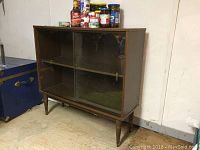Wood veneer cabinet with glass sliding doors, side view shows one fixed glass pane, legs, and stored items on top.
