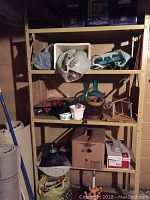 Three-tier shelving unit with various household and decor items including plant pots, wicker basket, candles, and cardboard boxes with extension cords.