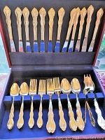 Photo of full open flatware box showing all utensils including knives, forks, spoons, serving pieces with gold plating and ornate handles arranged in blue felt compartments.
