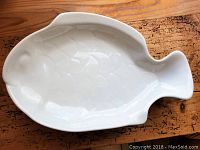 Single off white fish shaped ceramic plate shown on wood surface highlighting the shape and textured scale details.