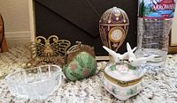 Five assorted trinkets including a gold-tone butterfly holder, small glass dish, green coin purse, purple egg-shaped box with clock face, and round floral box with doves, placed beside a water bottle for size reference.