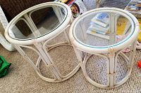 Two round rattan cocktail tables with clear glass tops, stacked together and placed on carpet flooring.
