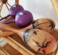 View of two pairs of maracas on a wicker shelf: one painted purple with Mexico and feather motifs, one natural wood with black painted floral and palm tree designs