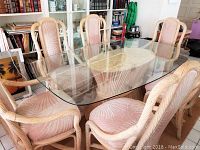 Full view of rectangular glass dining table surrounded by six upholstered cane/rattan chairs, showing clear temperate glass top and beige patterned upholstery with palm leaf motif.