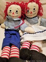 Full view of the front of two Raggedy Ann and Andy cloth dolls seated side by side on a beige surface