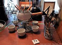 Photo of ceramic white fondue pot with wooden handle on metal burner stand, surrounded by 6 ceramic ramekins and 6 wooden-handled fondue forks inside the pot.