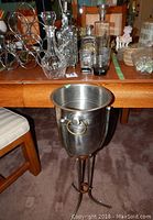Photo showing glass decanters, wire wine rack, shot glasses with wooden holder on table, and metal ice bucket on stand in foreground.