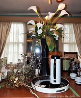 Photo of large black plastic vase with white faux flowers on dining table surrounded by glassware and other decor