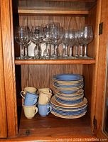 Wood cabinet shelf showing stacked stoneware dinnerware plates, bowls, and mugs in blue and yellow with 8 mugs total and 8 sets of plates and bowls.