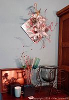 Two decorative square mirrors each 12x12 inches with floral pink arrangements on a blue wall, a framed embossed copper artwork, metal wine rack candle holder, six colorful champagne flutes, and two boxed mugs arranged on wood furniture.
