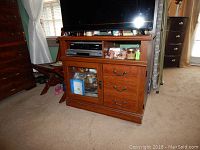 Front view of wooden TV chest holding a flat screen TV, a DVD player, and various small items on open shelf below TV. Cupboard with glass door and three drawers to the right visible.