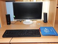 Black Dell monitor on beige wood shelf, with two black cylindrical Dell speakers on either side and a black Dell wired keyboard in front.