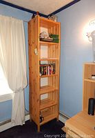 Tall, narrow wooden bookcase against blue wall by window with white curtains. Shelves hold books and decorative items including boxes and small figurines.