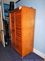 Wooden armoire photographed from an angle showing the left side and front with two sliding shingled doors on wheels.