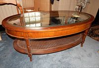 Oval glass coffee table with wooden frame and woven rattan lower shelf, showing tape residue and scuffs on glass top.