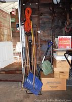 Photo showing a group of yard tools including shovels, a rake, and a broom standing against a wall inside a garage space.