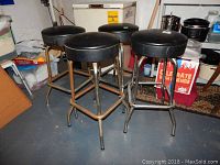 Six metal bar stools with black vinyl padded seats arranged in a cluster on a concrete floor, surrounded by storage boxes and household items.