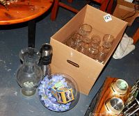 Photo shows a cardboard box with approximately a dozen clear stemware glasses inside, assorted types. Also visible are a glass pitcher and other items outside the box.