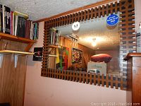 Front view of the large rectangular wall mirror with the carved wooden interlocking frame hanging on a wall with bookshelves above and adjacent furniture.