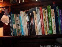 Wide view of the cookbooks tightly packed on a shelf, showing a mix of spiral bound and hard/soft cover editions, titles visible including Bread, Baking, Foreign Foods, Amish Cooking, and Official Cookbook.