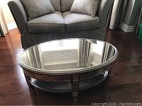 Angled side view of the oval cocktail table showing the mirrored top and lower shelf with brushed bronze wood frame and detailing