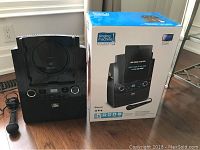 Black Singing Machine Classic karaoke player with lid open, showing CD compartment area, control panel, and front speaker. Wired microphone is lying on the floor. Original product box is placed next to the device, showing product image and features.