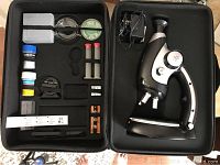 Open black hard case showing EduScience microscope and included accessories such as slides, petri dishes, lenses, and tools.