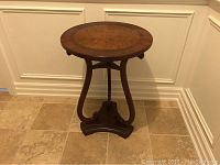 Photo of a small round Bombay end table with wooden top, curved legs, and triangular base, placed against a wall with white paneling.