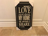 Photo shows the wall art still with protective corner covers, leaning against a white wall on a tile floor. The phrase displayed is clearly legible.