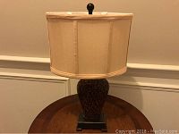 Lamp on a round wooden table showing textured brown and beige body with fabric drum shade and wood base.