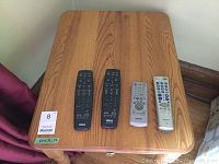 Top view of one wooden occasional table showing wood grain surface with four remote controls placed on top for scale comparison, sticker showing dimensions and lot number visible.
