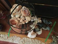 Photo showing metal basket filled with assorted small seashells on floor next to larger seashells and duck figurines.