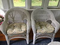 Front view of two white wicker chairs with floral seat cushions on a wooden floor in front of windows.