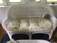 Front angled view of white wicker love seat with floral padded cushion showing seat, armrests, and woven backrest.