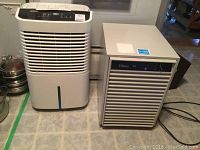 Two dehumidifiers side by side on a tiled floor, one white Whirlpool and one beige Classic D15.