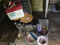 Lot showing cooler on top of wooden stool, second wooden stool adjacent, black folding chair, purple plastic jug and tray with colorful plastic glasses, melamine plates and floral plate on floor