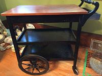 Full side view of black wooden serving cart with two black shelves, large wooden spoke wheels, and wooden top.