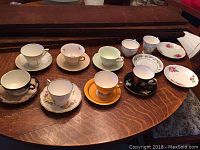 Full view of 8 cups and matching saucers laid out on a wooden table showing various styles and patterns.