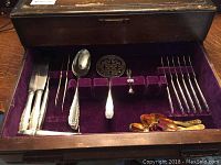 Wooden box with purple velvet lining containing knives, forks, spoons, serving utensils, and gold tone teaspoons organized in compartments.