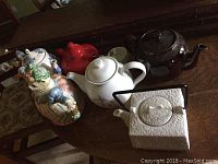 Collection of seven assorted teapots displayed on wood table, includes Fitz and Floyd cat teapot, Corelle blue iris white teapot, red teapot with chip, brown Brown Betty, white Sadler with chips, Asian Morris Design teapot, and white textured square teapot with black handle.