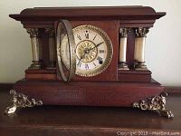 Front view of the antique Beth Thomas mantle clock showing the dial with Roman numerals, brass center, wooden case with decorative columns and brass feet.