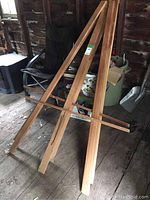 First image showing one wooden easel leaning against a structure inside a shed or garage with visible floorboards.